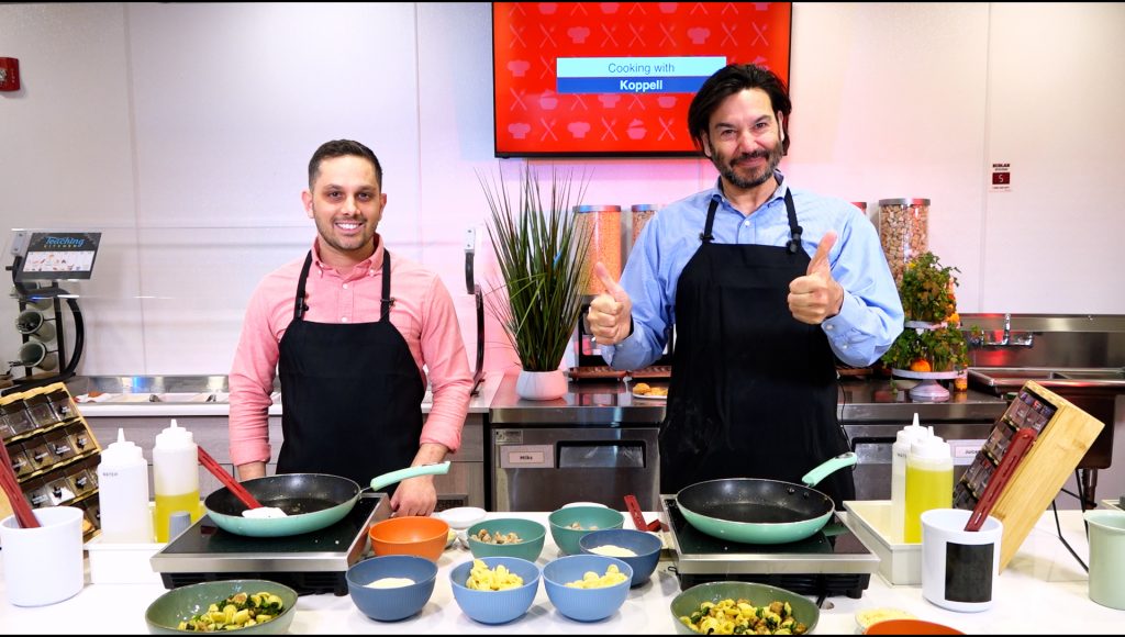 Two men standing in front of a stove both smiling and standing. The man on the left has a long-sleeved pink button down shirt and the man on the right is giving a "thumbs up" sign and wearing a long-sleeved button down blue shirt.