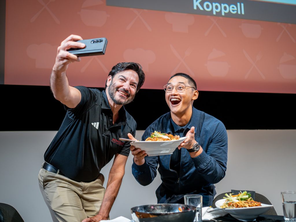 Two men posing for a "selfie" photo, one on the left dressed in a black polo shirt holding a mobile phone, and the other holding a dish of food. Both are smiling and happy.