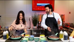 A woman on the left and a man on the right are standing in front of a stove preparing a meal. Each are tending to separate pans to help prepare the dish.