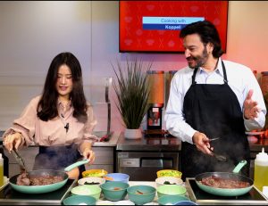 A woman on the left and a man on the right are standing in front of a stove preparing a meal. Each are tending to separate pans to help prepare the dish.