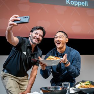 Two men posing for a "selfie" photo, one on the left dressed in a black polo shirt holding a mobile phone, and the other holding a dish of food. Both are smiling and happy.