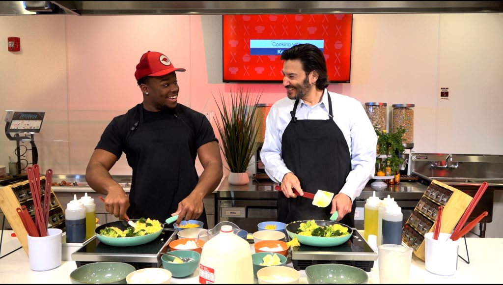 Two men standing in front of a stove preparing a meal. The man on the left is wearing a black shit and red hat and the man on the right is wearing a long-sleeved button down white shirt.